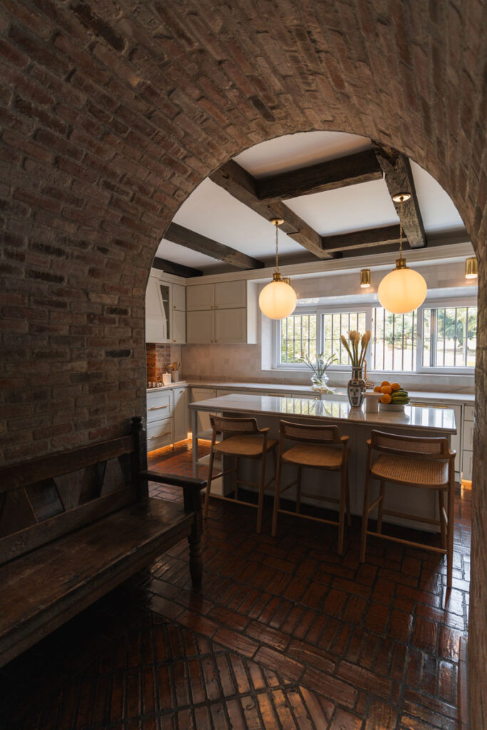 Cocina en madera clara con cubierta blanca, fregadero tipo granja y ventanal de doble altura sobre la estufa a gas.